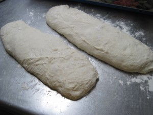 once ciabatta dough has proofed for a 2nd time, use 2 dough scrapers and pick up the dough from either end and turn upside down onto a paper lined and flour dusted sheet pan